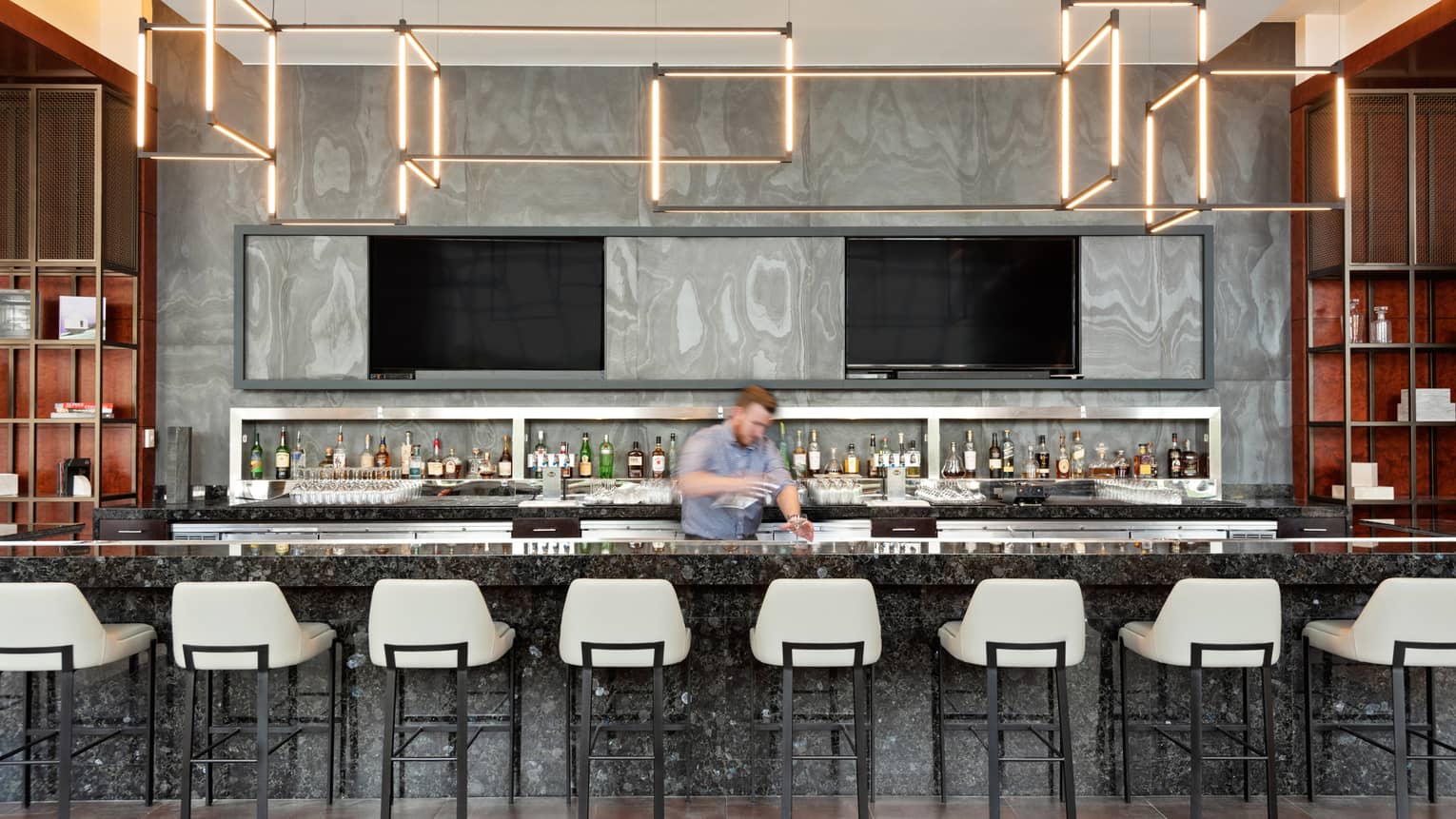 A bartender working behind a bar with white stools, a variety of liquor can be seen behind them.