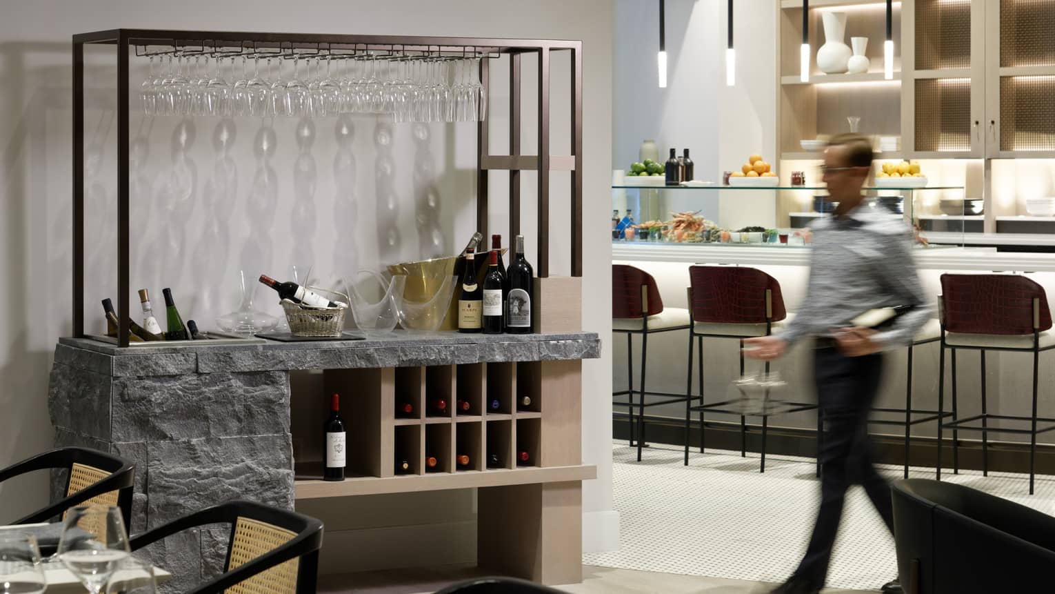 A staff member walking towards a shelf with wine and wine glasses on it.
