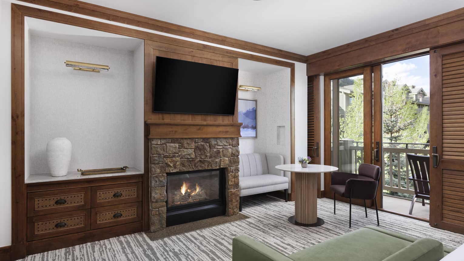 Luxury hotel room lounge area with a stone fireplace, mounted TV, green chaise, sofa and chair by the table, and a balcony with tree views