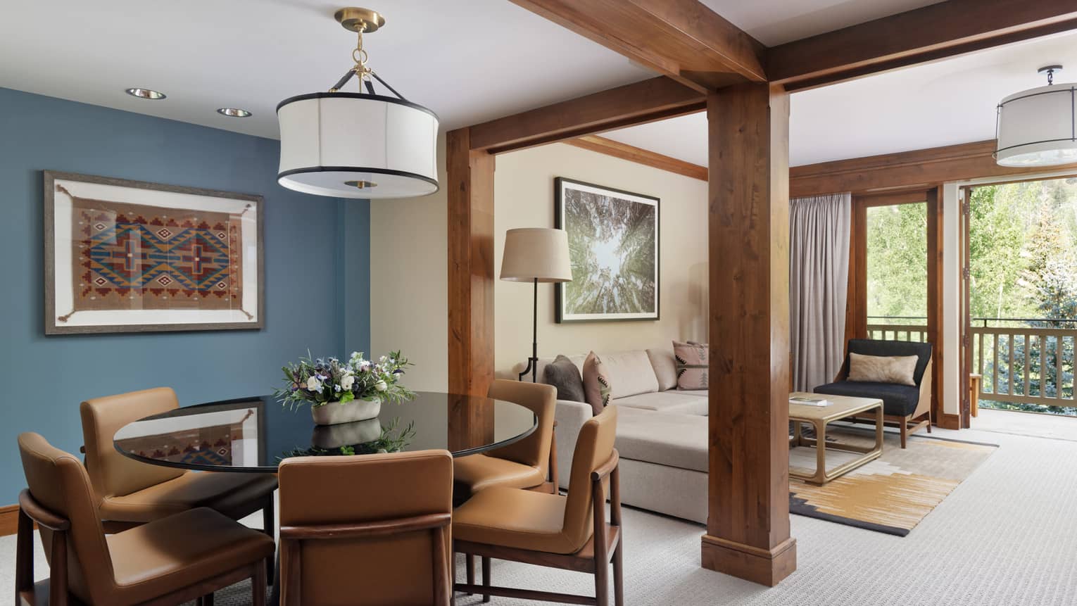 Dining area with a round table, leather chairs, and artwork on a blue accent wall, opening to a living area with a sectional sofa and balcony