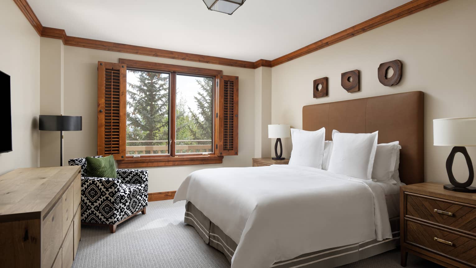 Mountain chic resort guest room featuring king bed with brown headboard, wooden-framed window with shutters, patterned arm chair and dresser