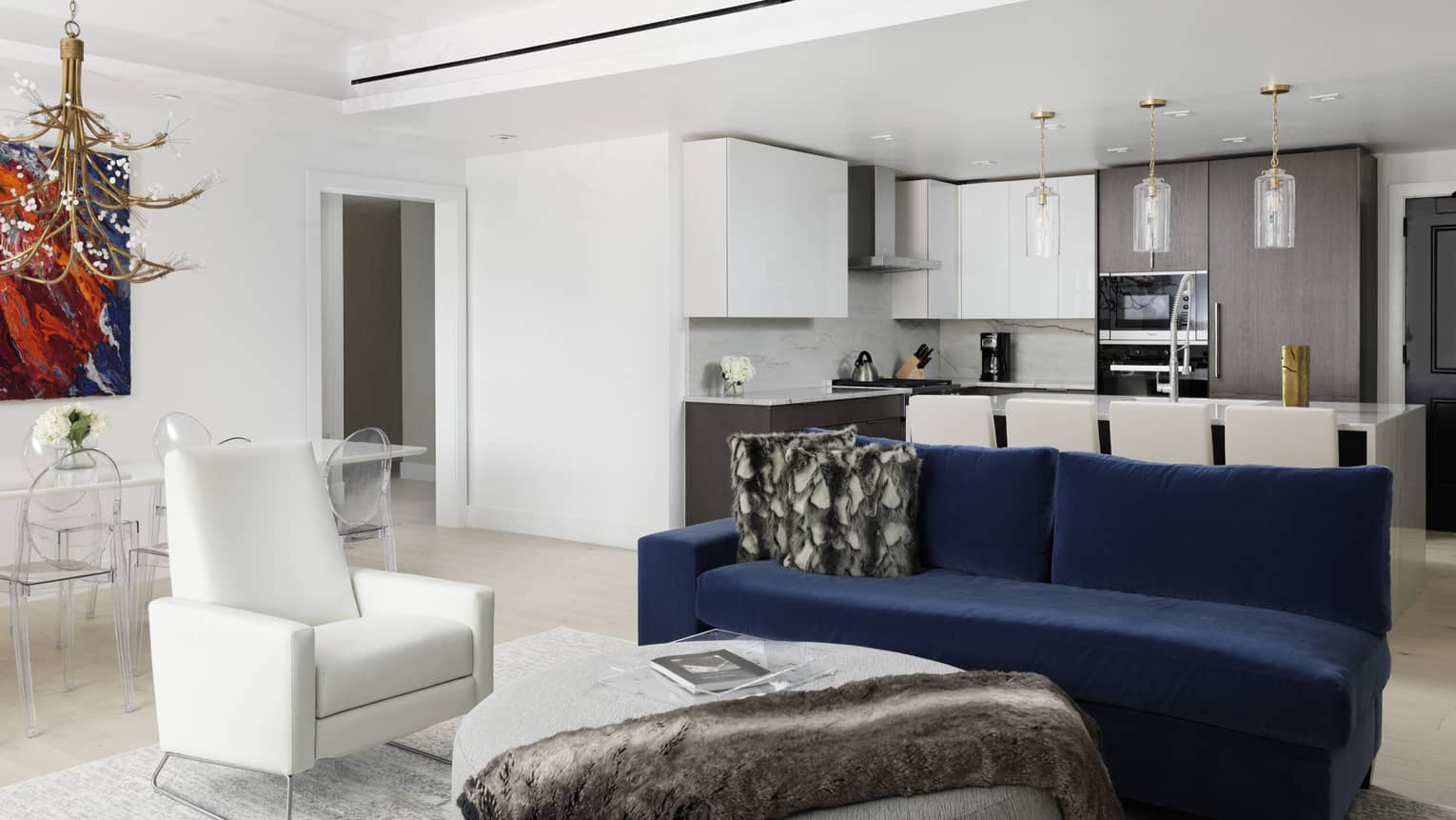 Living room with white walls and ceiling, dark blue sofa, white chair, kitchen in background