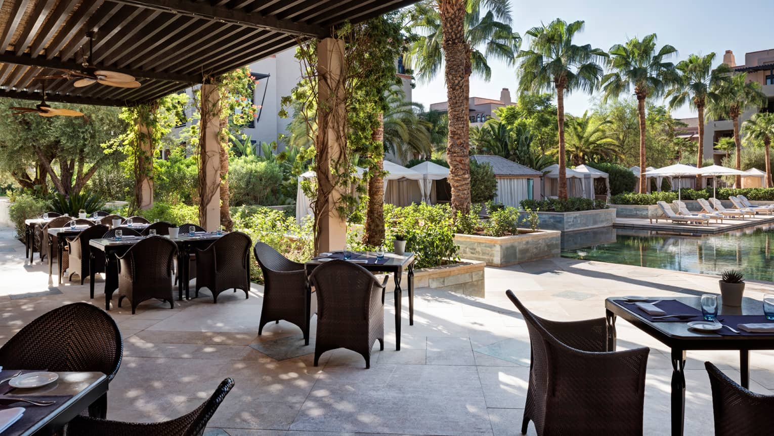 Patio tables and chairs under wood pergola near outdoor swimming pool, white cabanas 