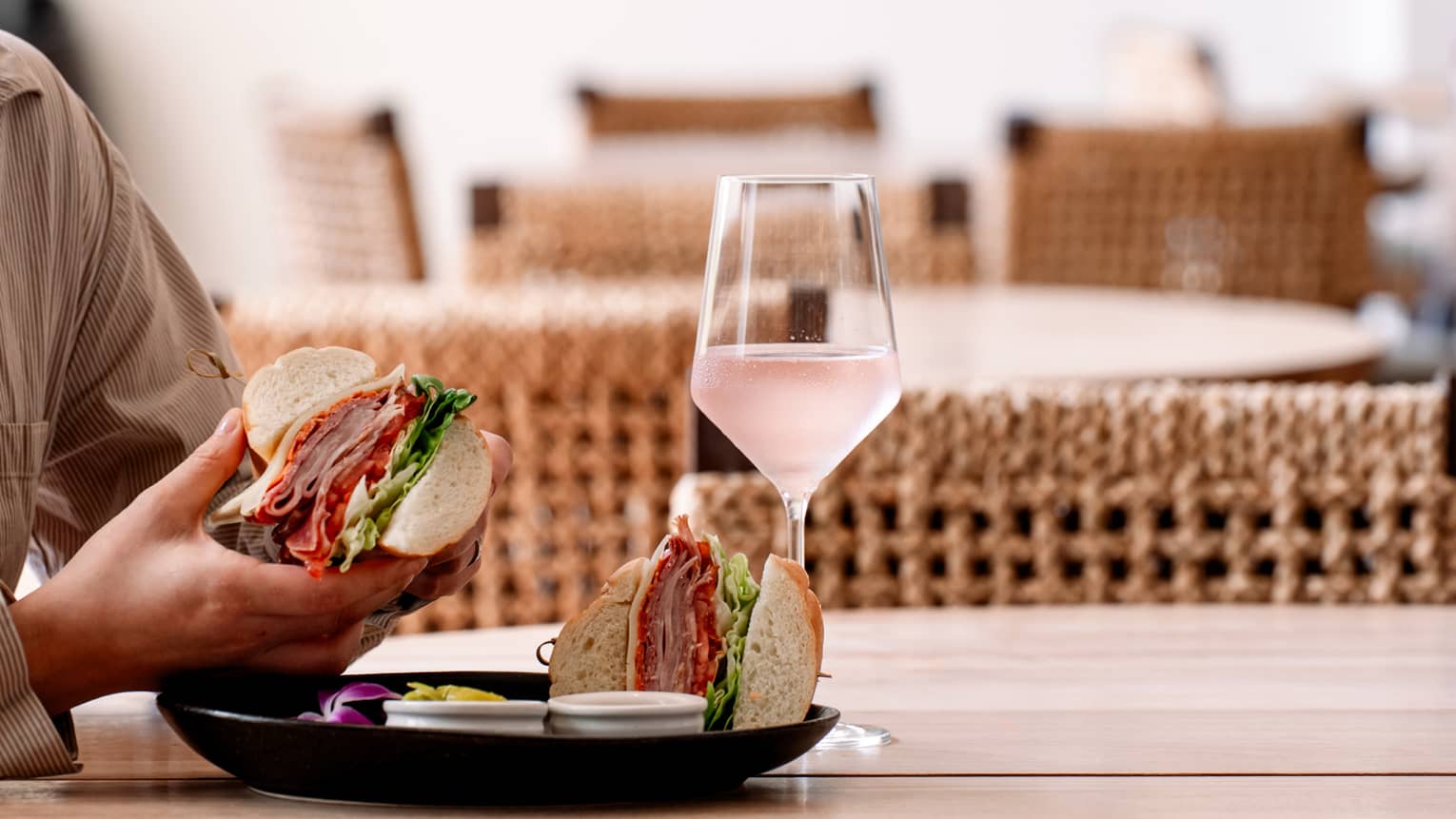 A person holds a large sandwich over a black plate on a wooden table, next to a glass of ros�