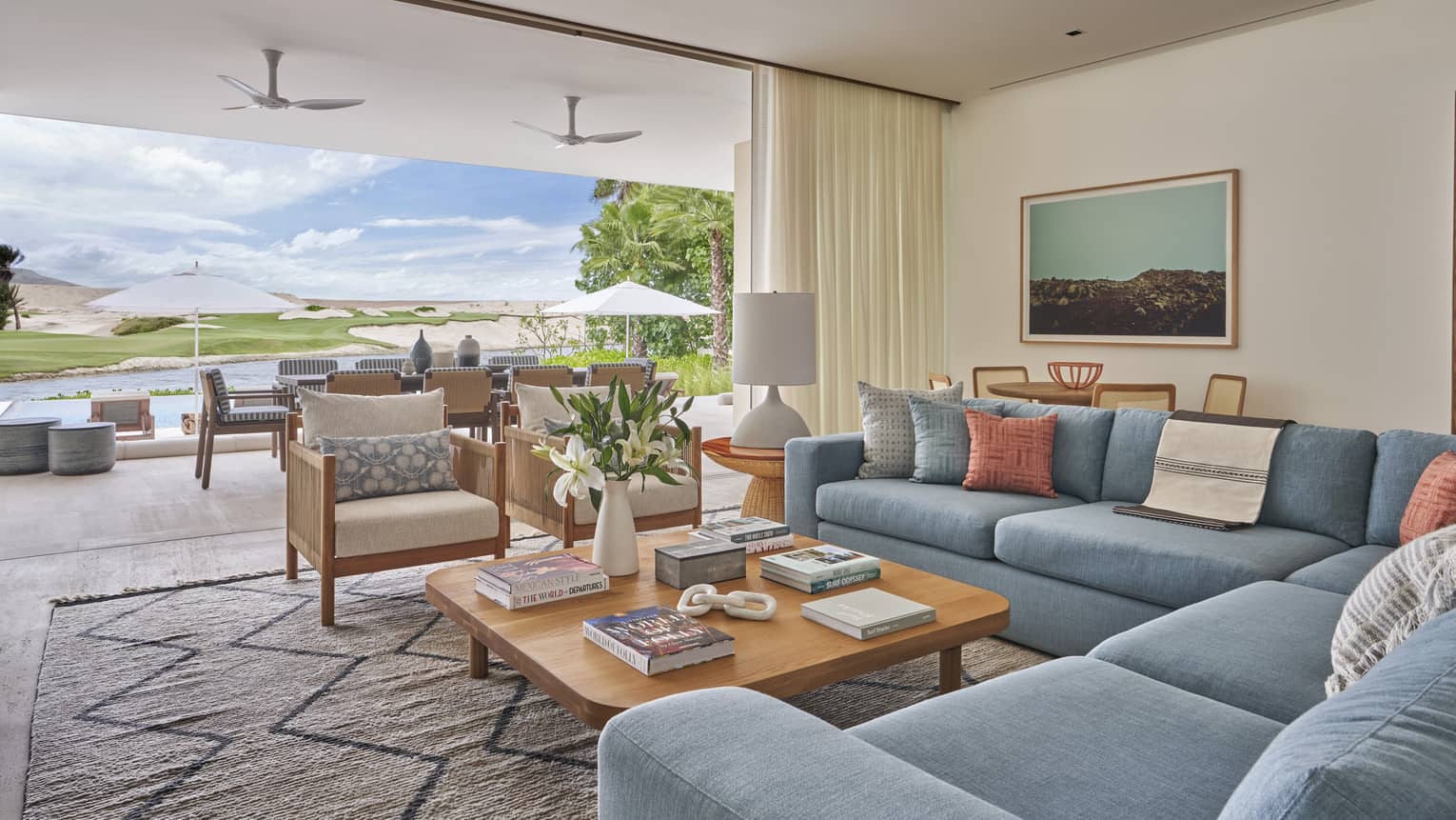Large modern living room with blue sectional, square wooden coffee table and two club chairs with wall open to terrace and golf course view