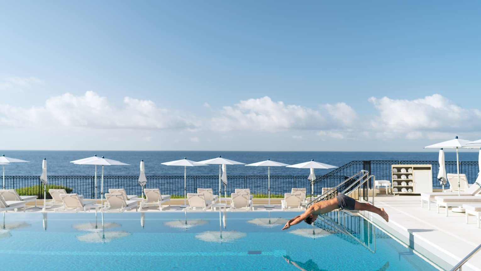 Man diving into blue Club Dauphin pool, white umbrellas and lounge chairs along edge, sea behind