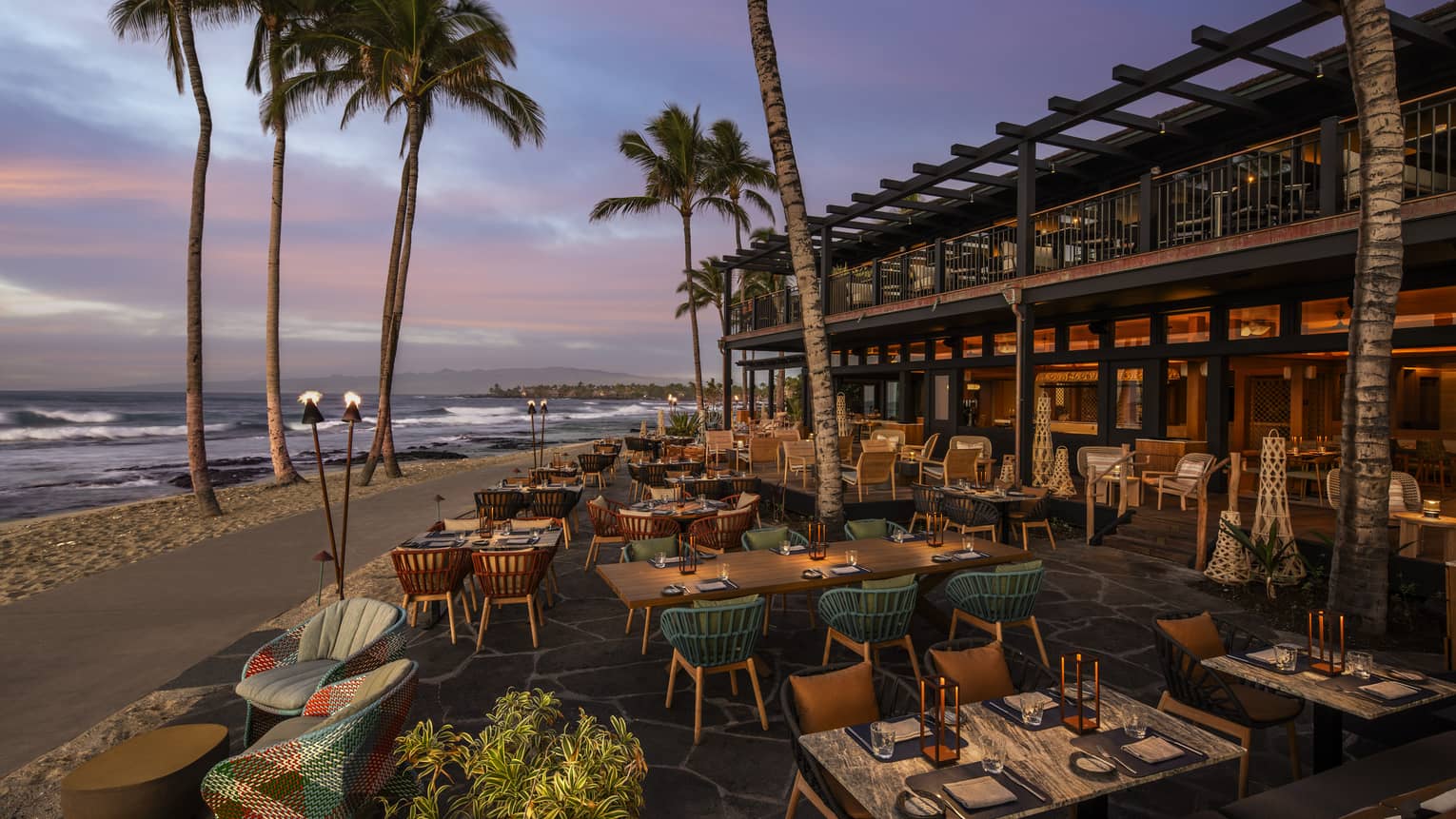 A beachside restaurant with a spacious outdoor dining area. In the background are palm trees, the ocean and the sky tinged with pink, grey, purple and blue.