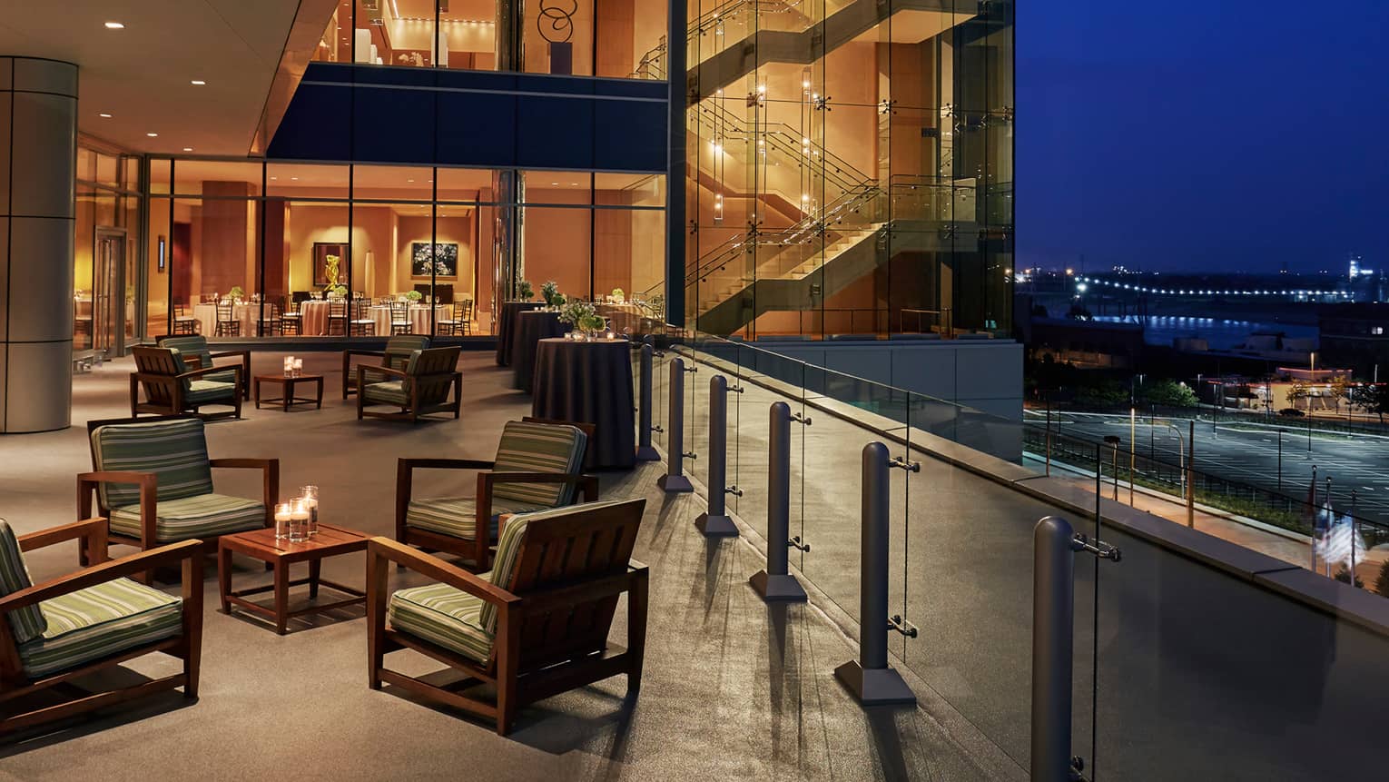 Lounge chairs around small cocktail tables with candles, near floor-to-ceiling glass window at dusk
