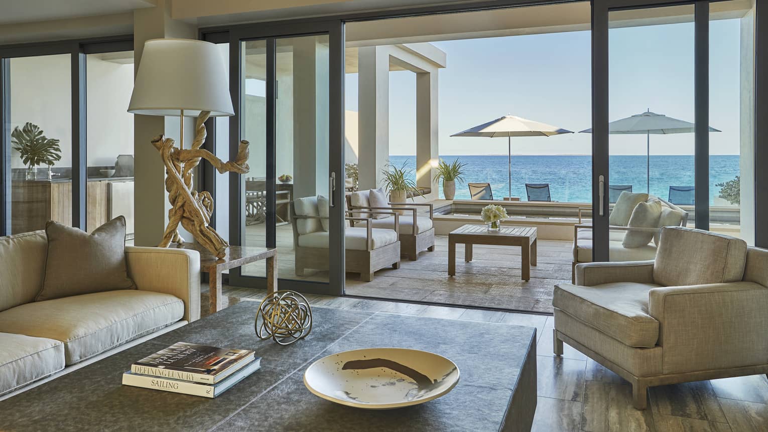 Elegant living room with neutral-toned seating, a coffee table and sliding glass doors opening to a terrace with ocean views and shaded loungers