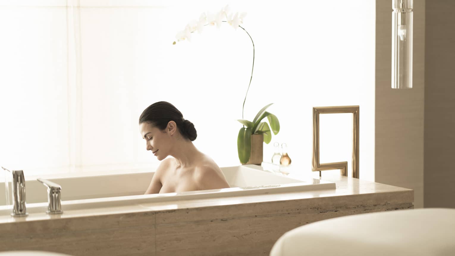 Dark-haired woman relaxes in bathtub in light-filled bathroom 