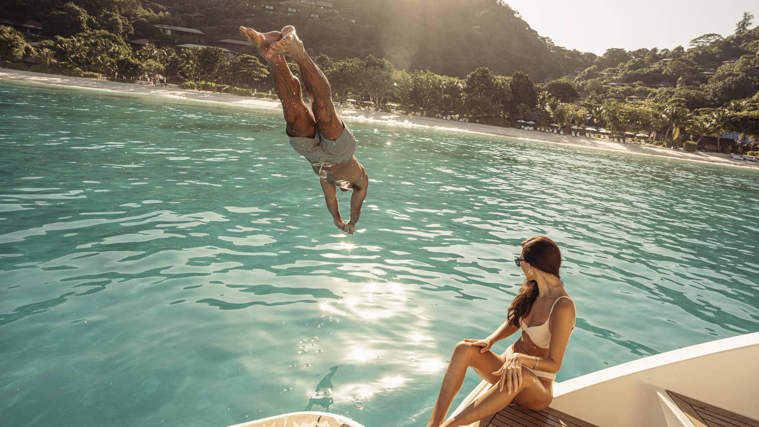 Person diving into blue-green water off a boat deck, where a person in swimsuit is sitting