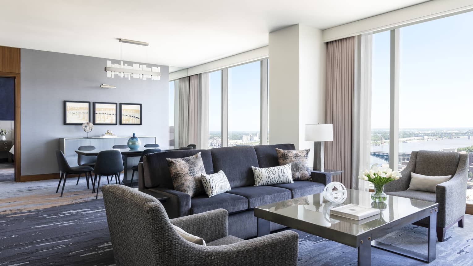 Hotel suite living room with dark blue sofa, two grey arm chairs, large square coffee table and wall of windows