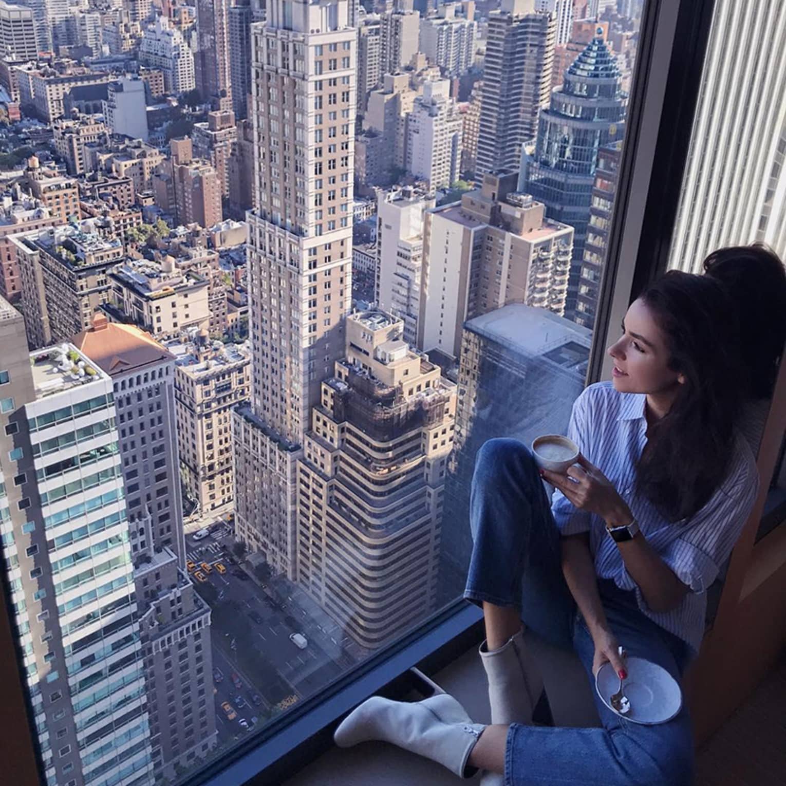 Woman with coffee cup sits in large window overlooking New York City rooftops
