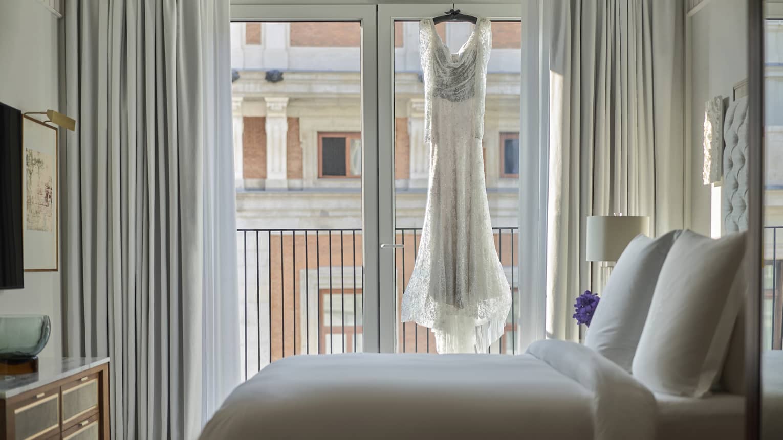A lace wedding dress hangs in front of a large window in a bright, elegant bedroom, with a view of a historic building across the street.
