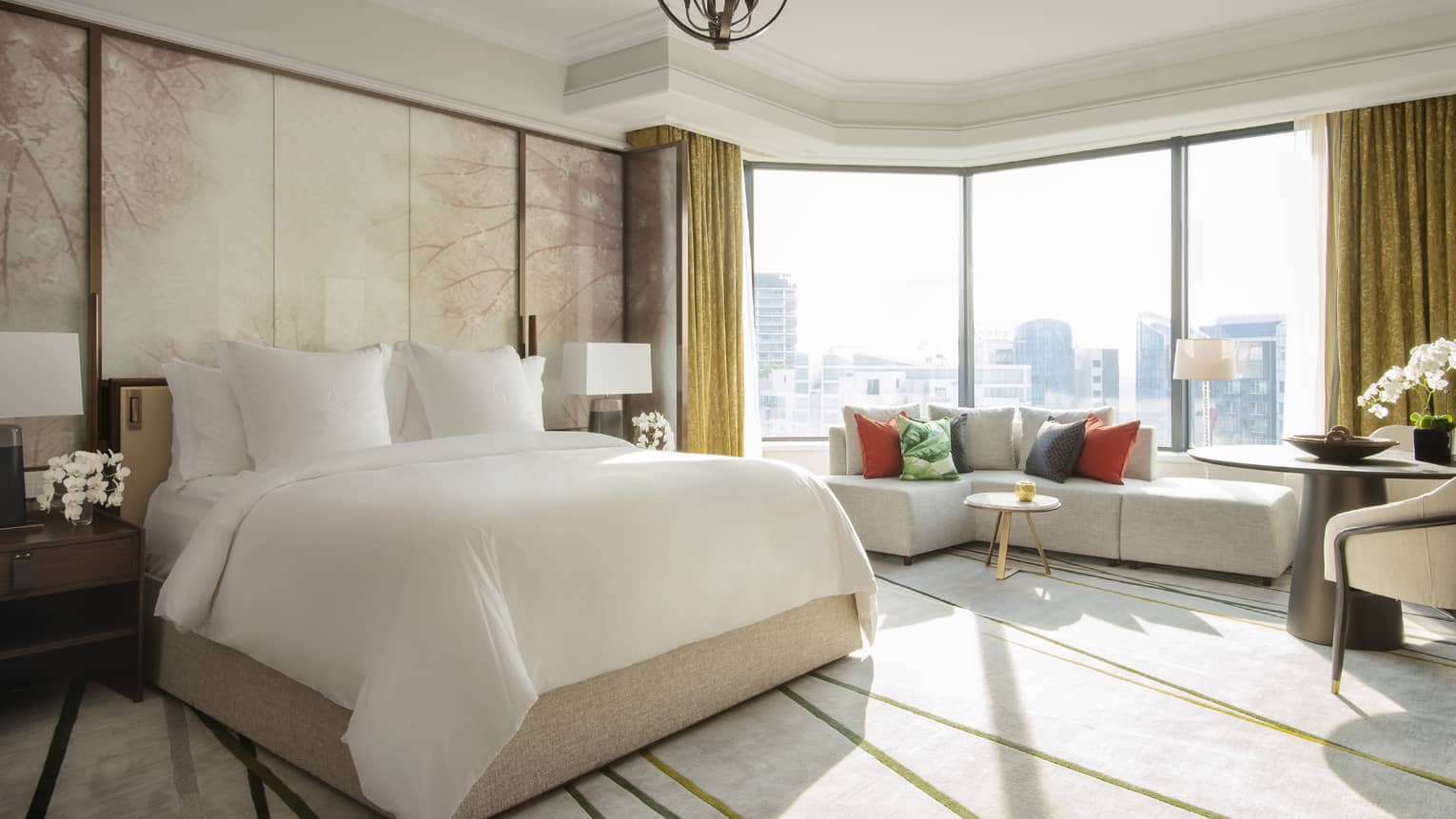 A spacious guest room featuring a large bed, seating area with colourful pillows and floor-to-ceiling windows with city views.