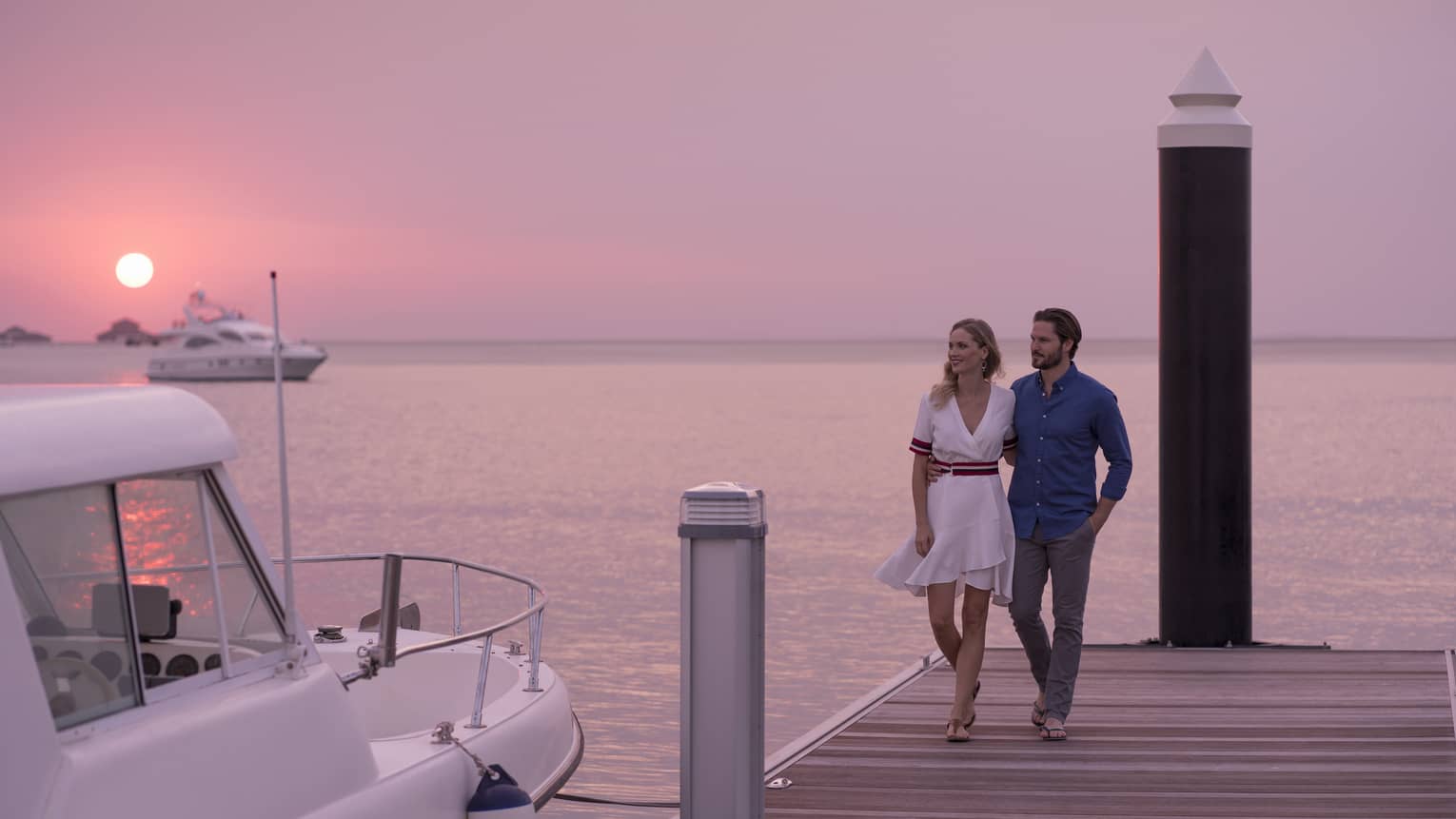 Couple walks across dock towards small yacht at sunset