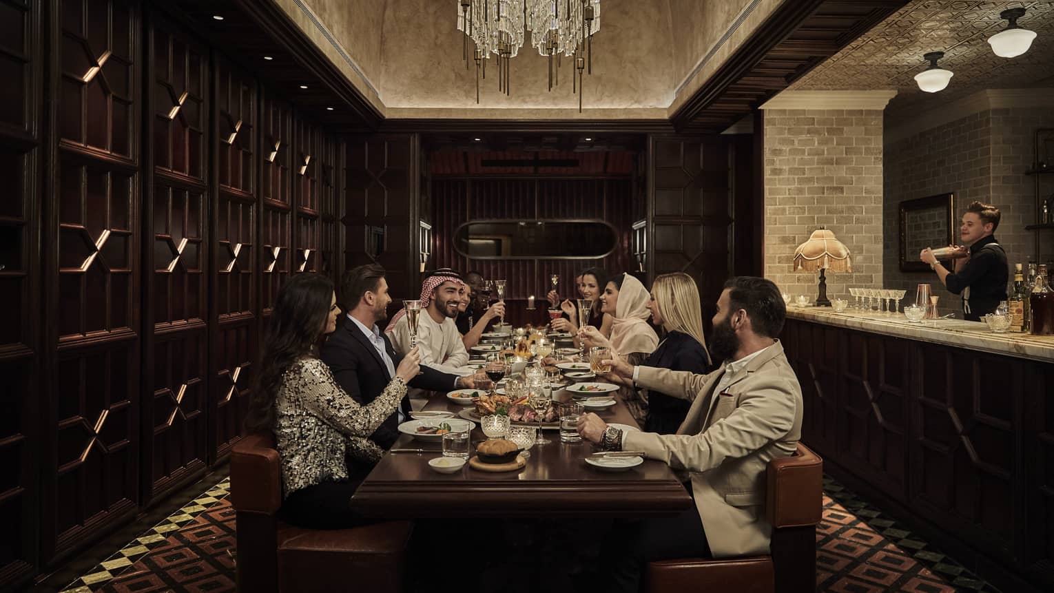 Ten people sit at a long, dark-wood rectangular table in a dark-wood panelled, speakeasy-style room with a crystal chandelier overhead and a marble bar counter and bartender to the side