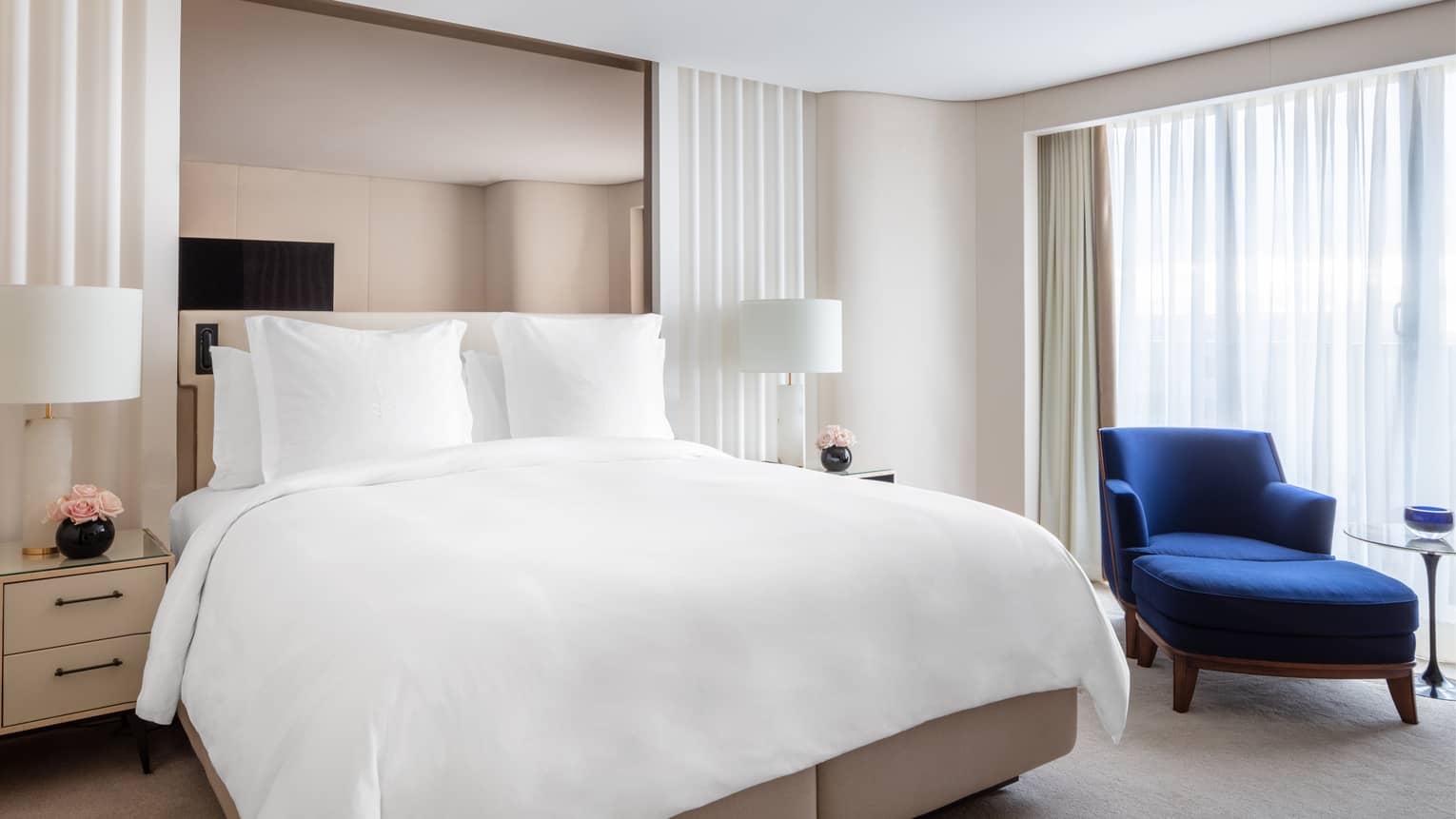 Bedroom with white king bed, royal blue chaise, mirrored headboard, white curtains, nightstands with pink flowers