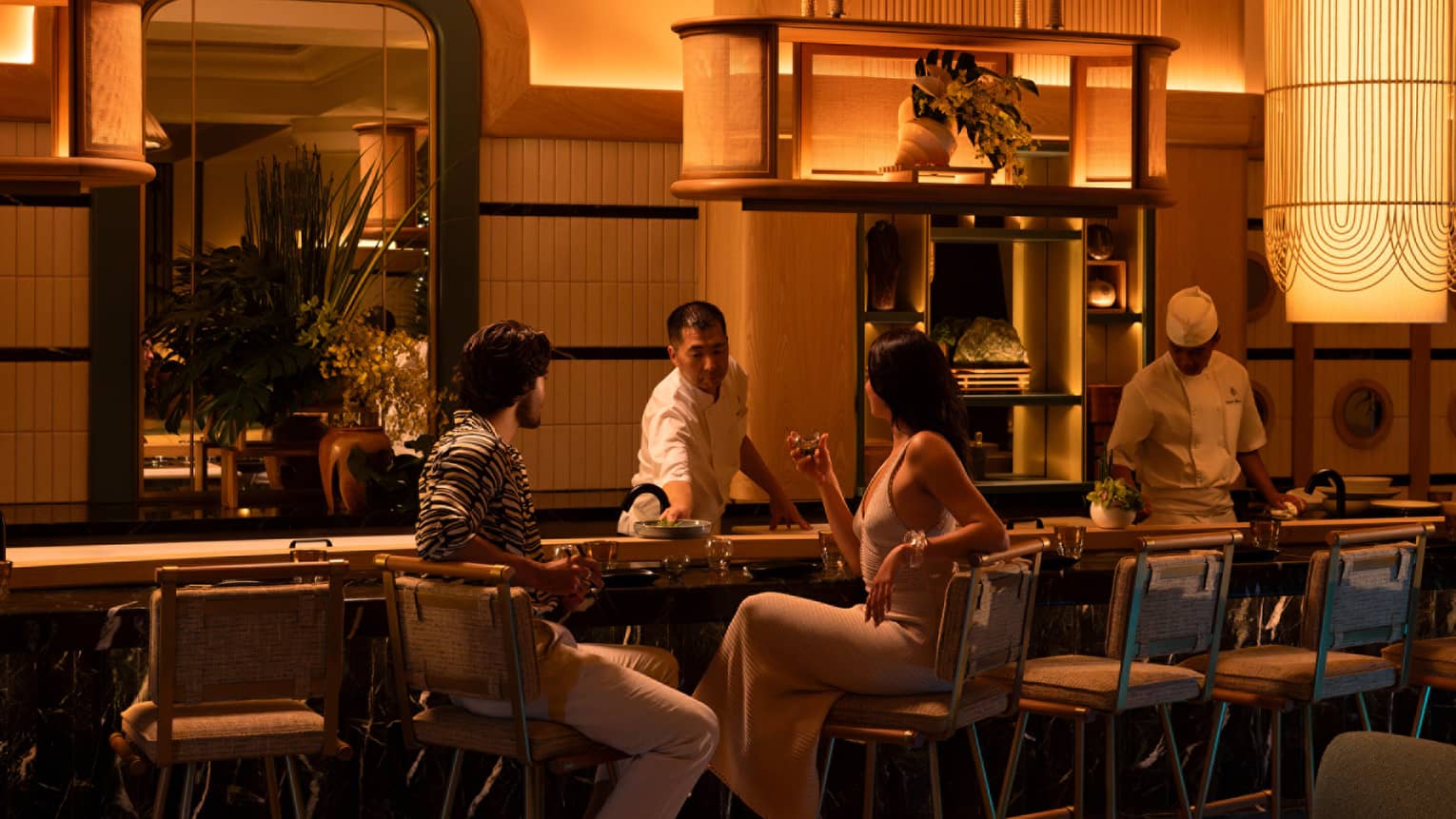 Two people sit at glamorous sushi counter at luxury hotel, dimly lit