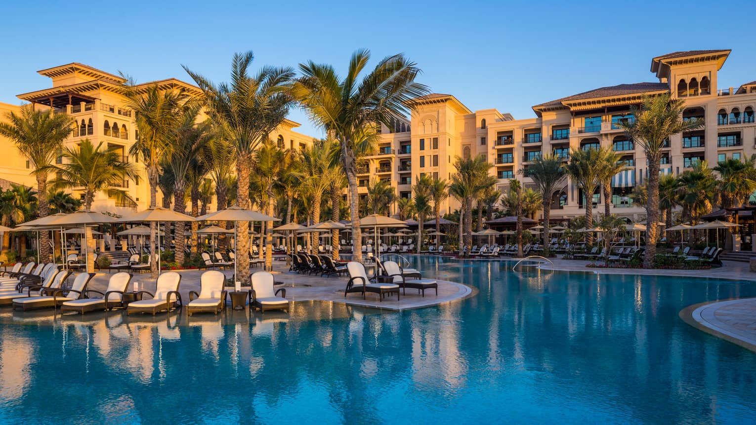Winding blue outdoor swimming pool lined with white chaise lounge patio chairs, palm trees