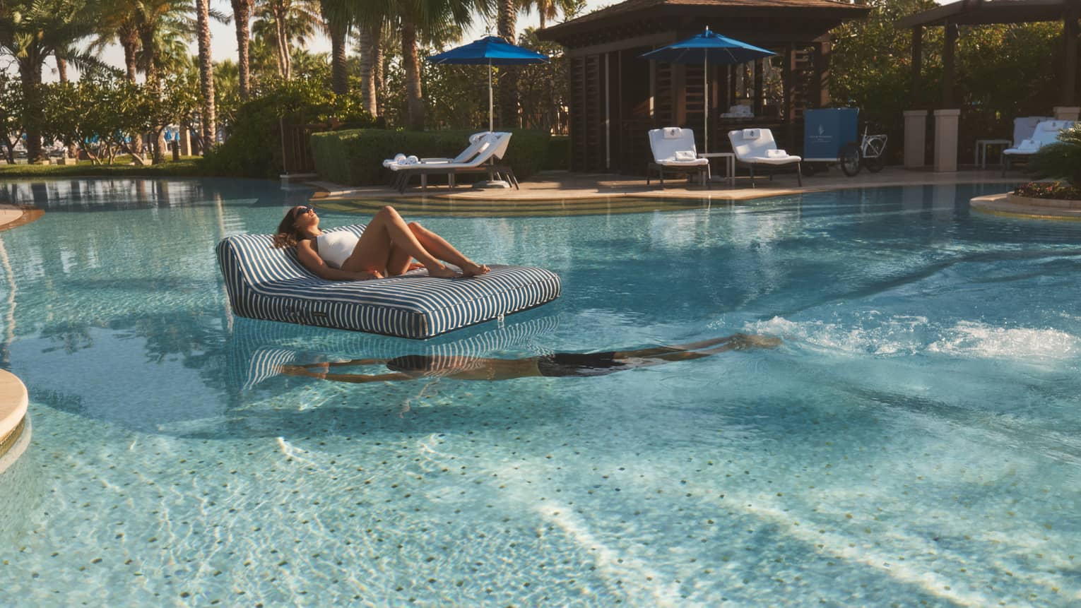 Person lounges on a black-and-white striped floating chair in the middle of a pool as another person swim beneath