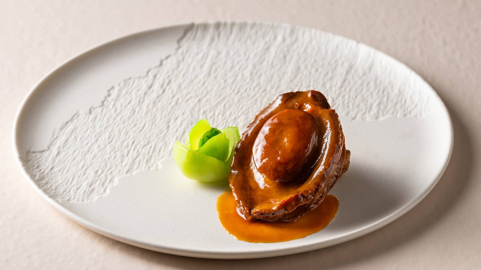 Textured white plate with abalone in sauce and green vegetable garnish