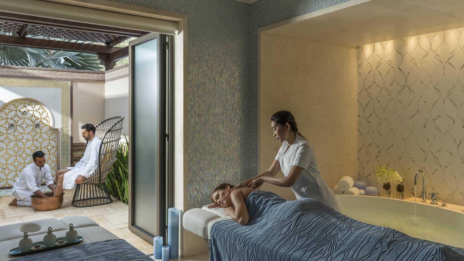 A woman under a blue sheet gets a massage from a masseuse surrounded by candles and natural light. 