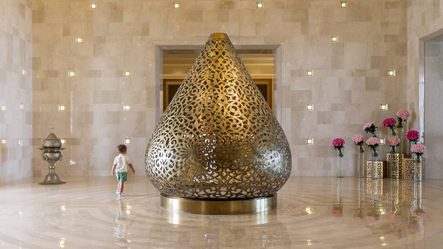 Child standing beside intricately carved gold conical sculpture in marble-lined hotel lobby