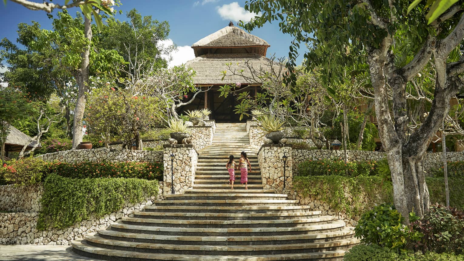 Back of two young girls walking up large stone staircase to stone lobby with thatched roof