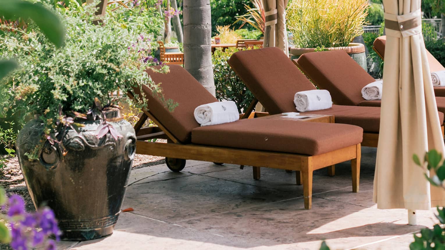 A poolside cabana with brown lounge chairs and plants and flowers surrounding it.