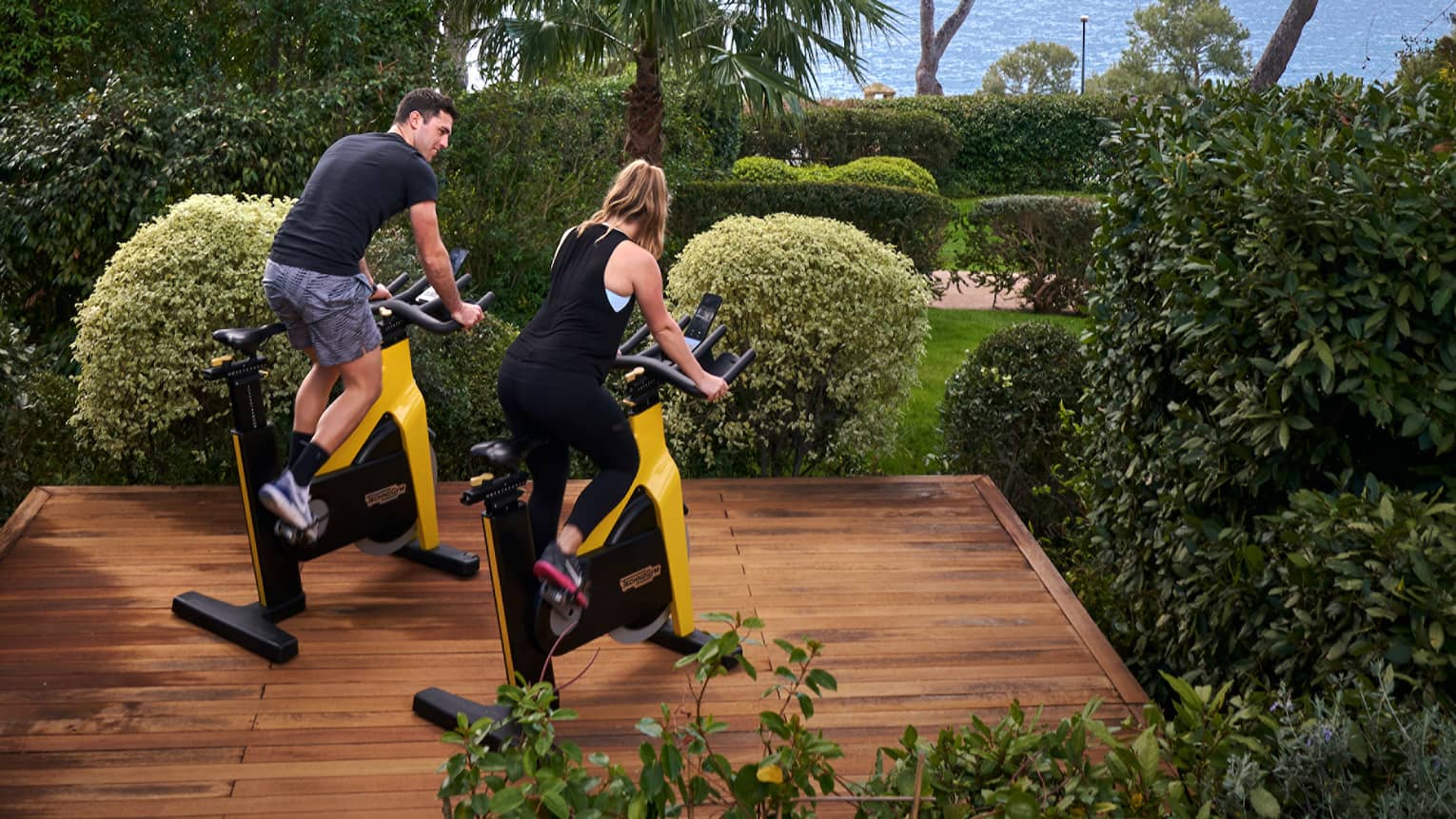 A man and a woman ride stationary bikes on outdoor wooden platform, sea in the distance