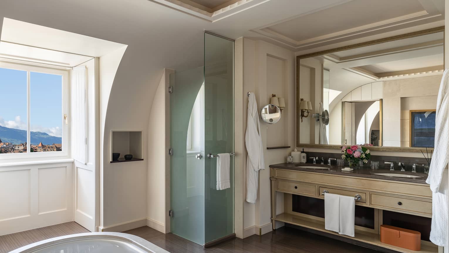 Luxury hotel bathroom with large mirror, double sink vanity, glass shower and soaking tub by the window offering city and mountain view