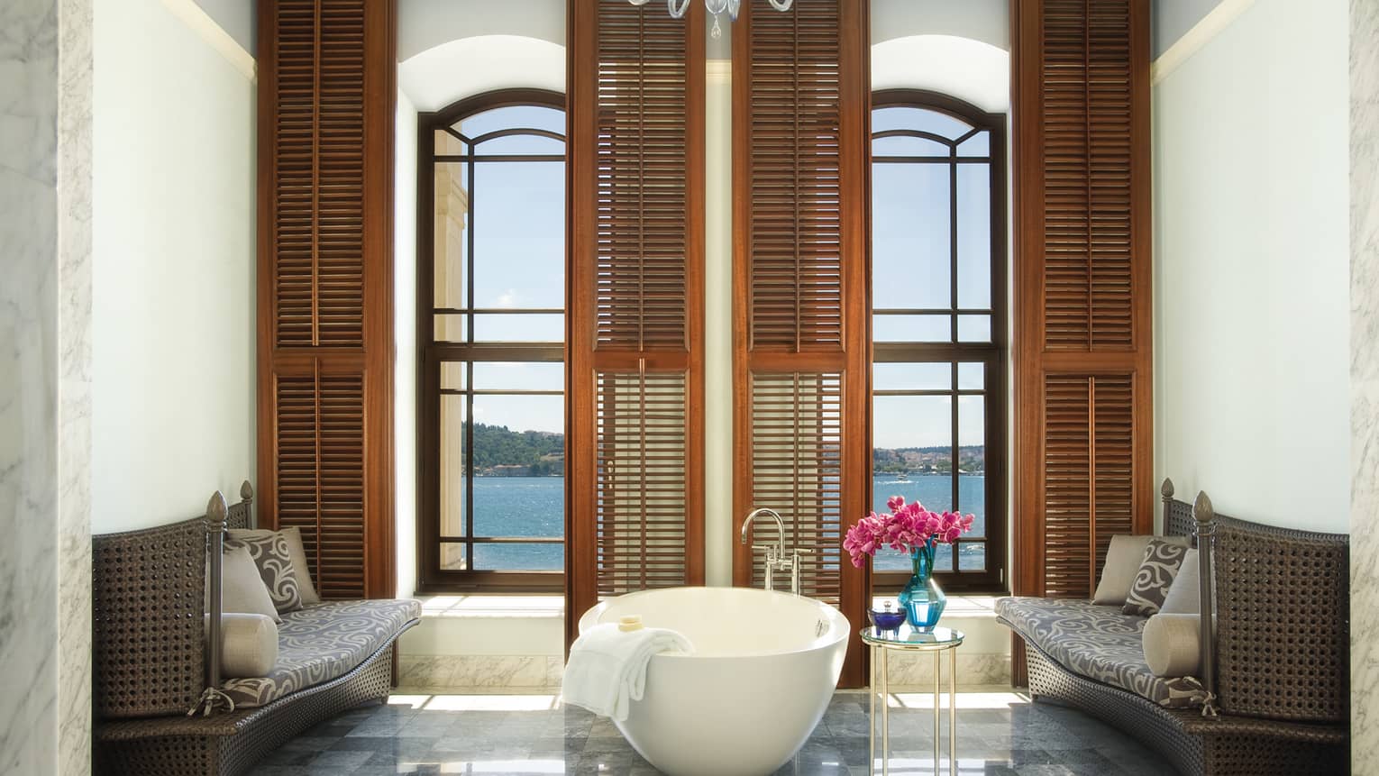 White freestanding tub in front of tall windows, wood shutters in marble bathroom