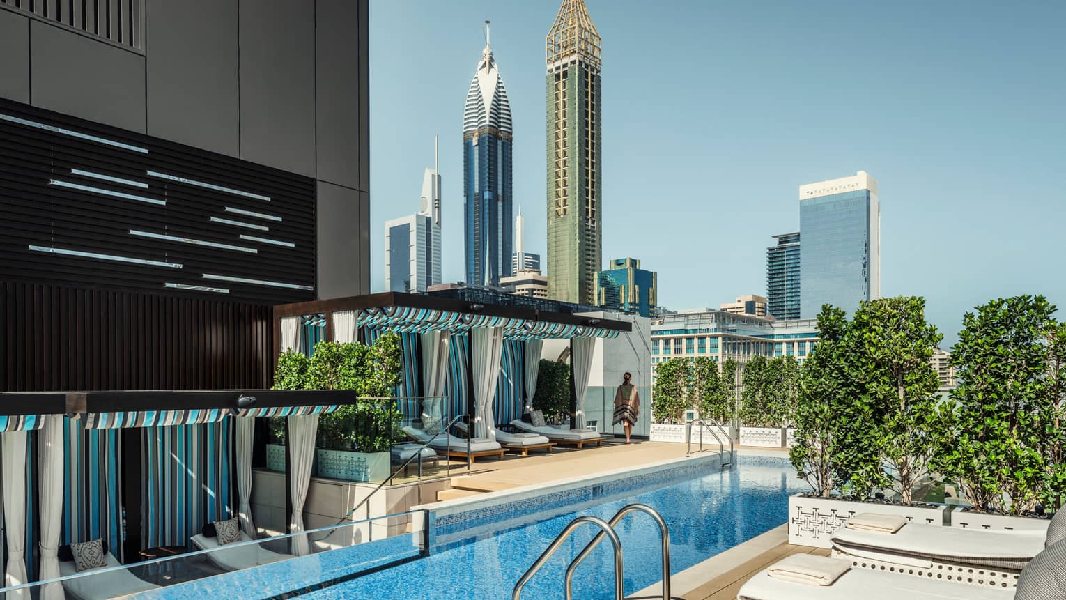 White lounge chairs, ladder into long blue swimming pool by cabanas on rooftop patio on sunny day