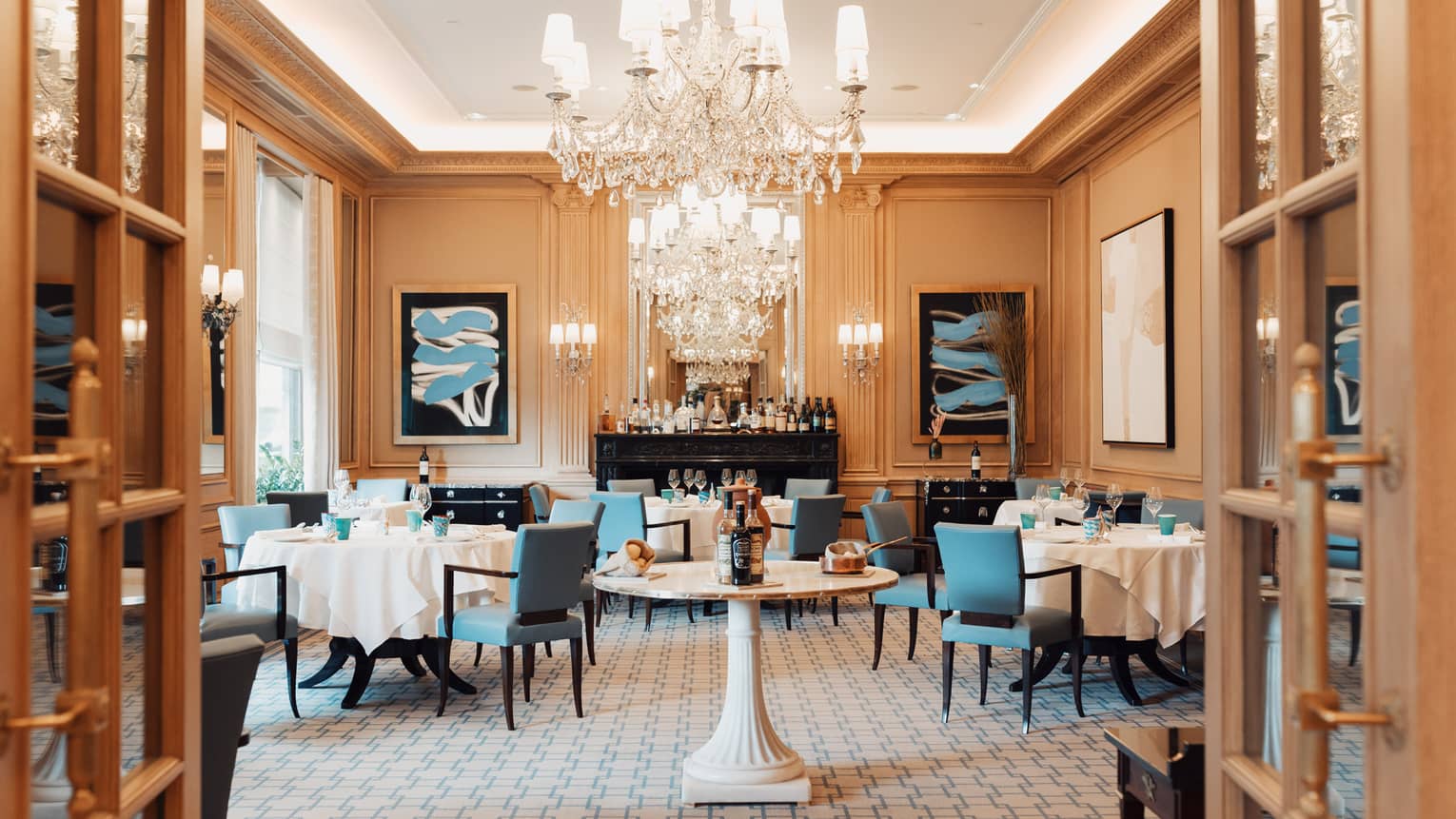 Elaborate crystal chandeliers sparkle in a cream, brown and blue-accented dining room containing tables set for a meal.