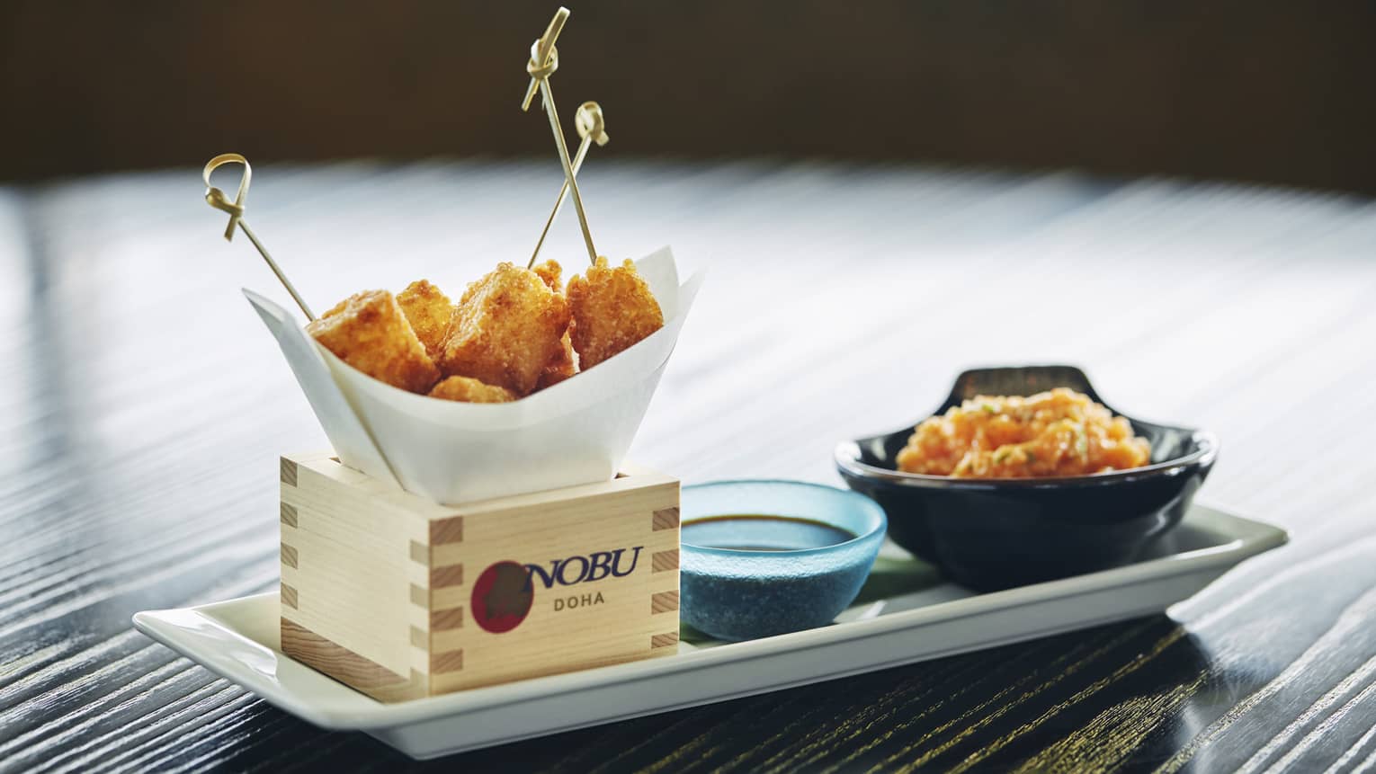 Close-up of white platter, wood box with Nobu logo, fried appetizers on toothpicks, dish with sauce