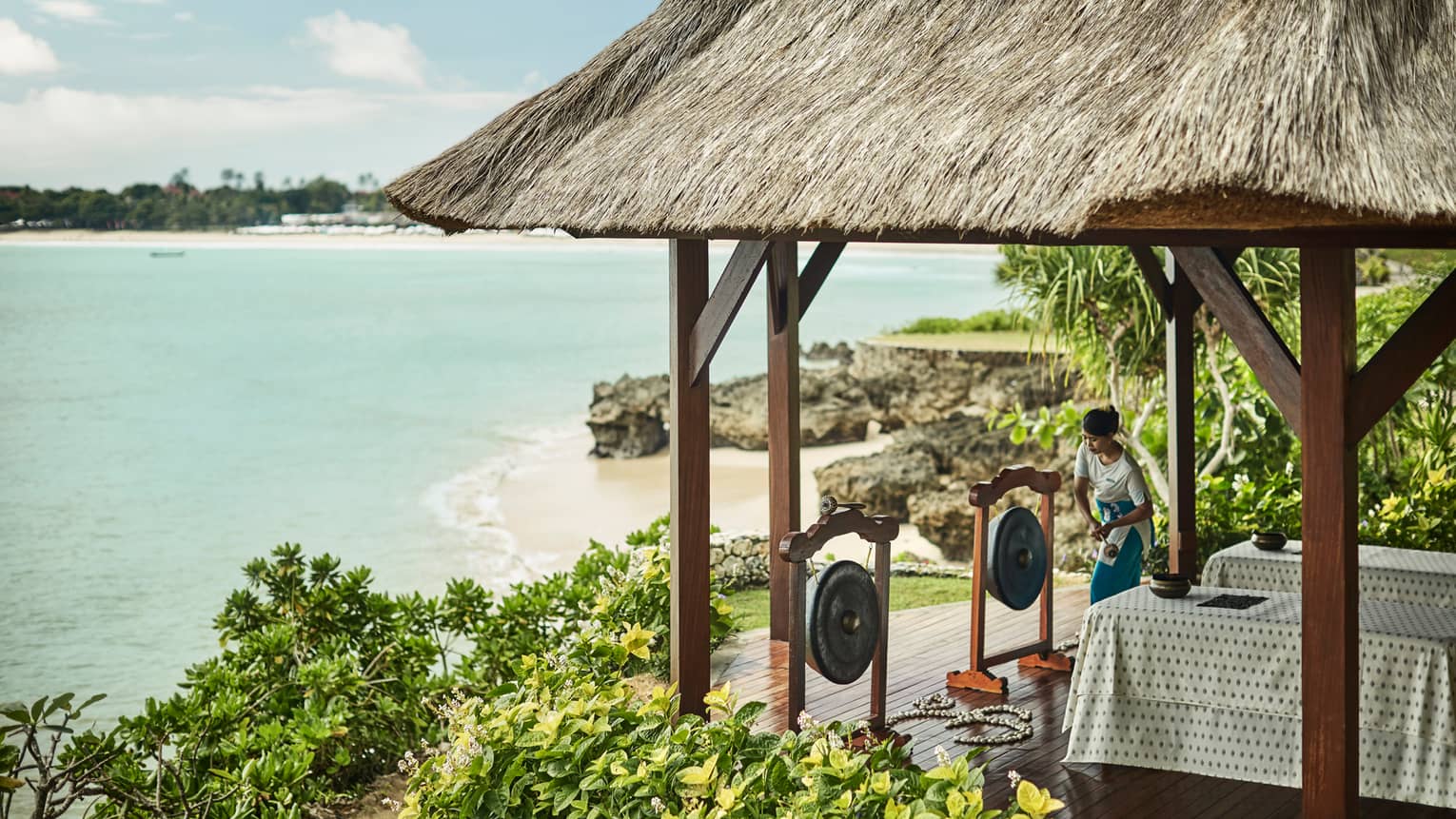 Spa attendant prepares to strike Balinese in front of two massage tables under gazebo overlooking beach, ocean