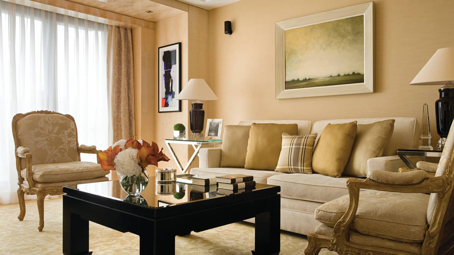 The Deluxe Suite at Four Seasons London Park Lane in Mayfair showcases a classic and luxurious living room setting. The room features a palette of soft beige and gold tones, with a plush beige sofa adorned with gold cushions. Two ornate, antique-style armchairs flank a sleek black coffee table, creating a sophisticated and inviting conversation area. Large windows are draped with heavy curtains, allowing natural light to enhance the warm atmosphere. The decor is completed by a traditional landscape painting above the sofa and elegant table lamps.