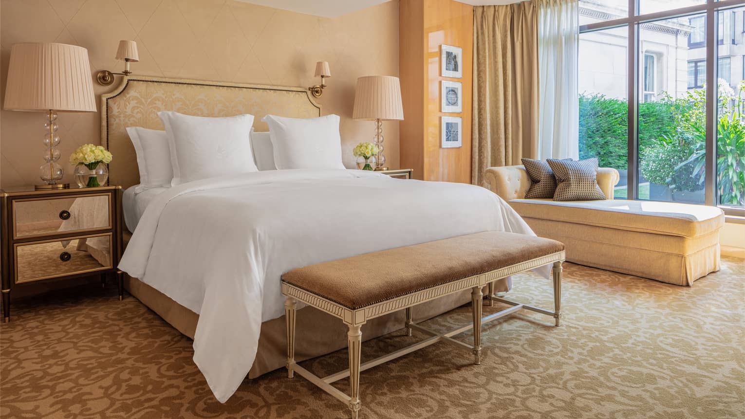 White bed with tan bench at foot against tan wall and chaise longue by large window