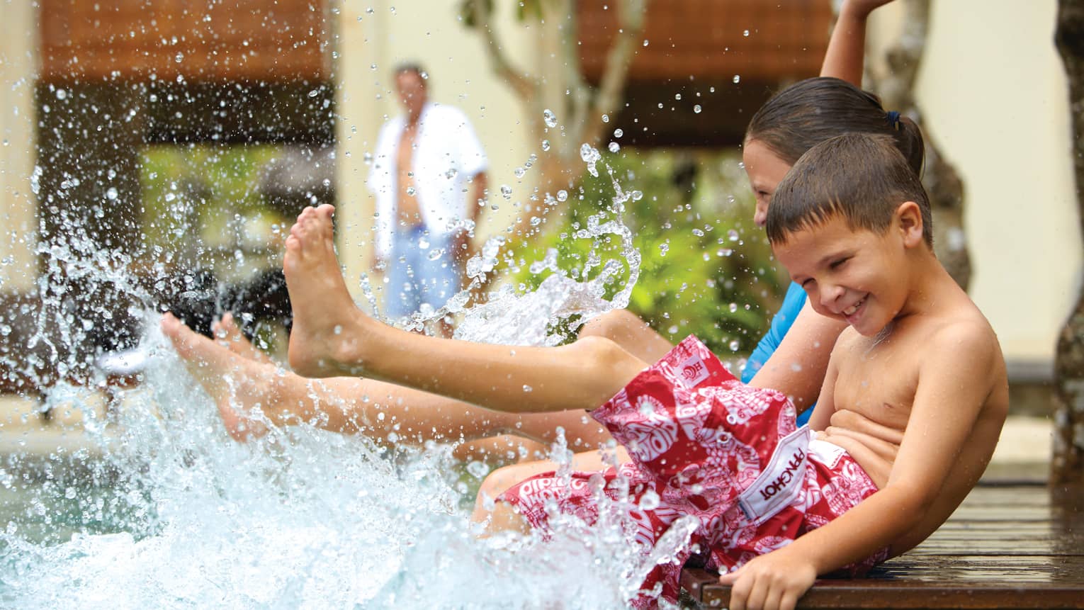 Young children in swimsuits sit on edge of swimming pool, splash water with legs