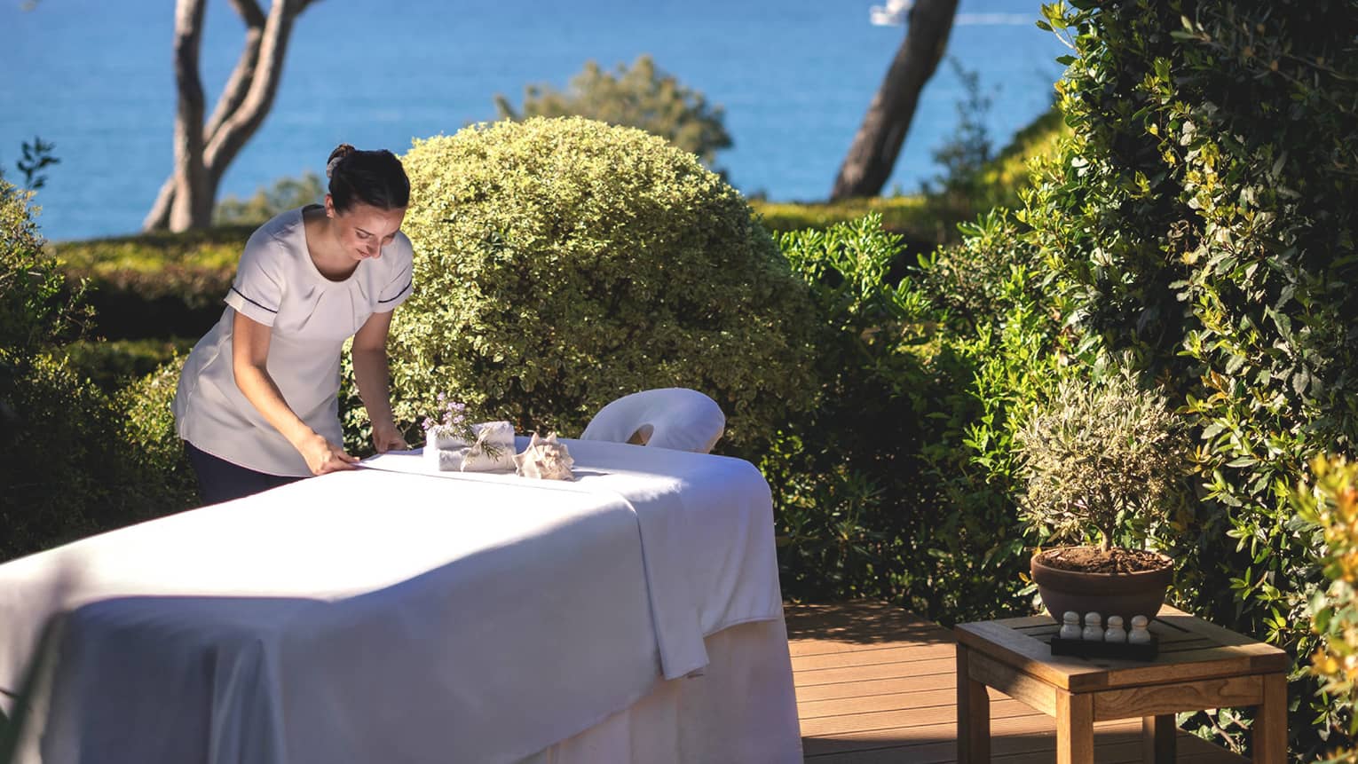 Spa therapist setting up spa table on outdoor platform surrounded by bushes and trees and overlooking the sea