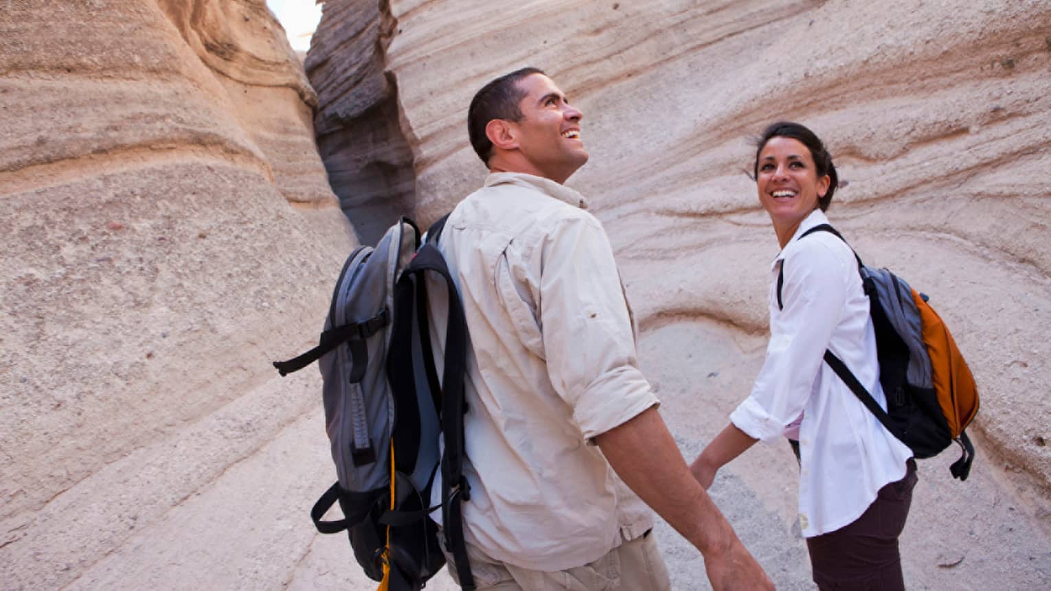 Smiling man and woman wearing backpacks hold hands, walk through desert rocks