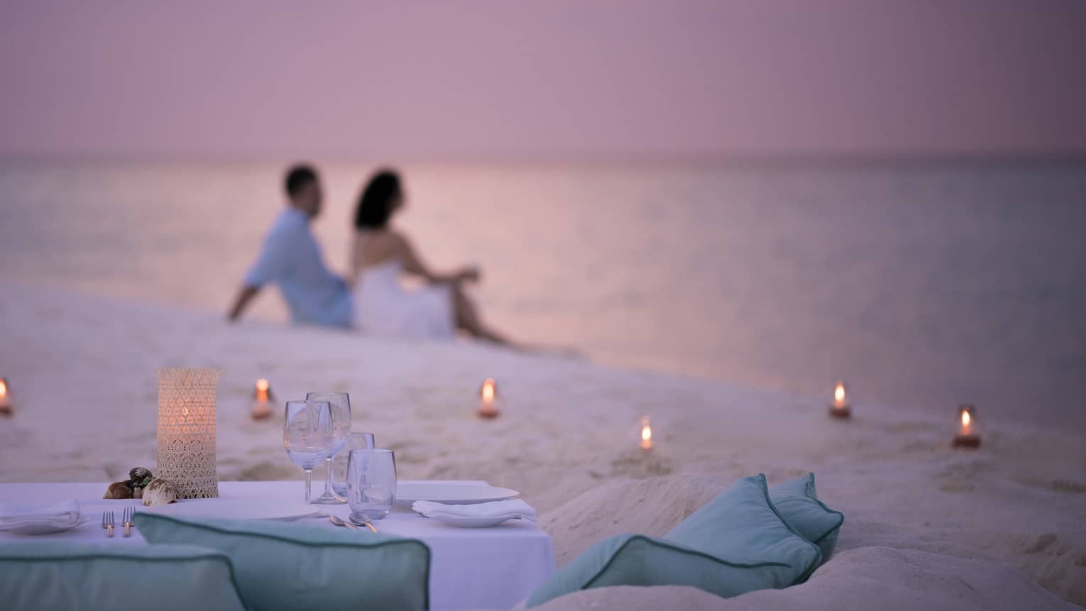 Private dining table in front of sand beach with tealight candles, couple sitting in sand