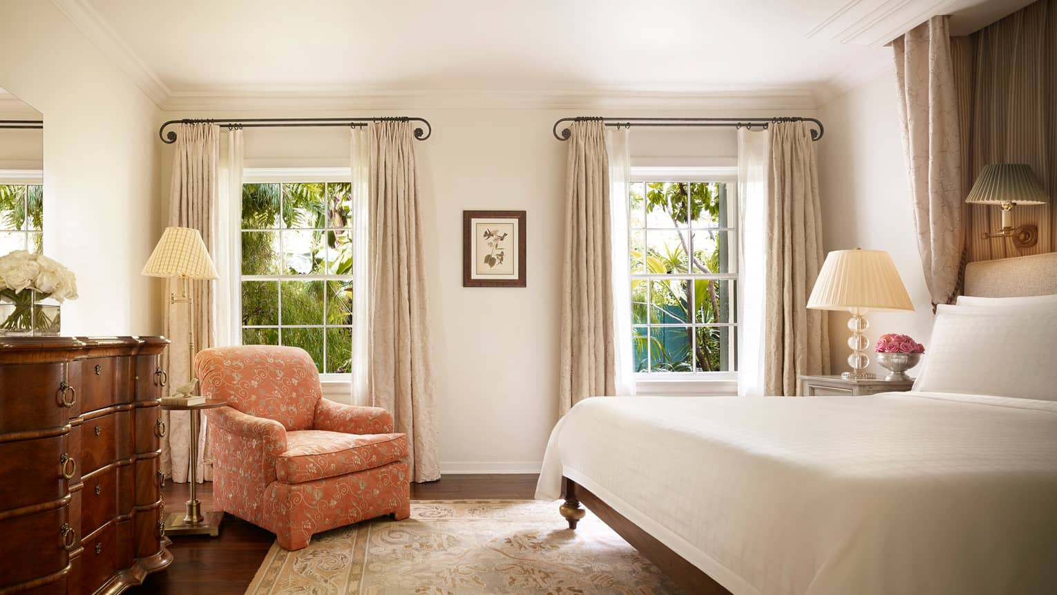 Wood dresser, pink floral armchair by tall sunny windows, nightstand and bed