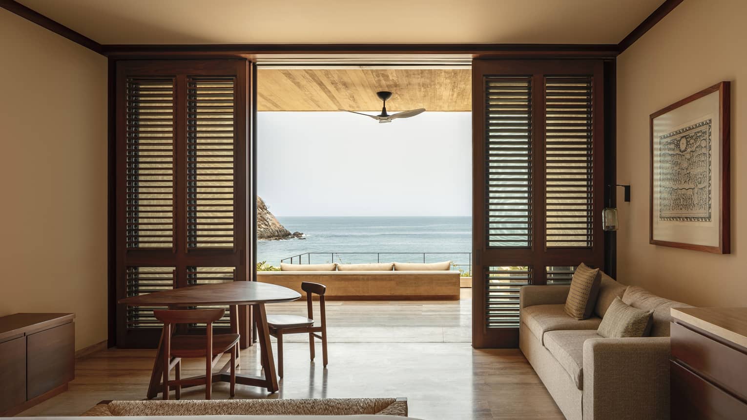 Bedroom with wooden shutters opening to a terrace with ocean views. The room features a cozy sofa, a small dining table and a ceiling fan on the patio.