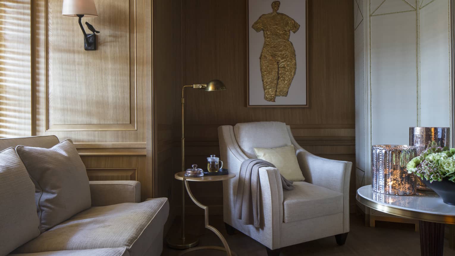 Sunlight streams through window to corner of sofa, white armchair with blanket on arm, reading lamp, art piece on wall