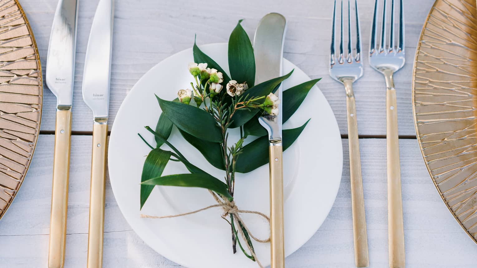 A bohemian table setting of gold chargers, modern long gold and silver flatware, white porcelain plates and a sprig of greenery