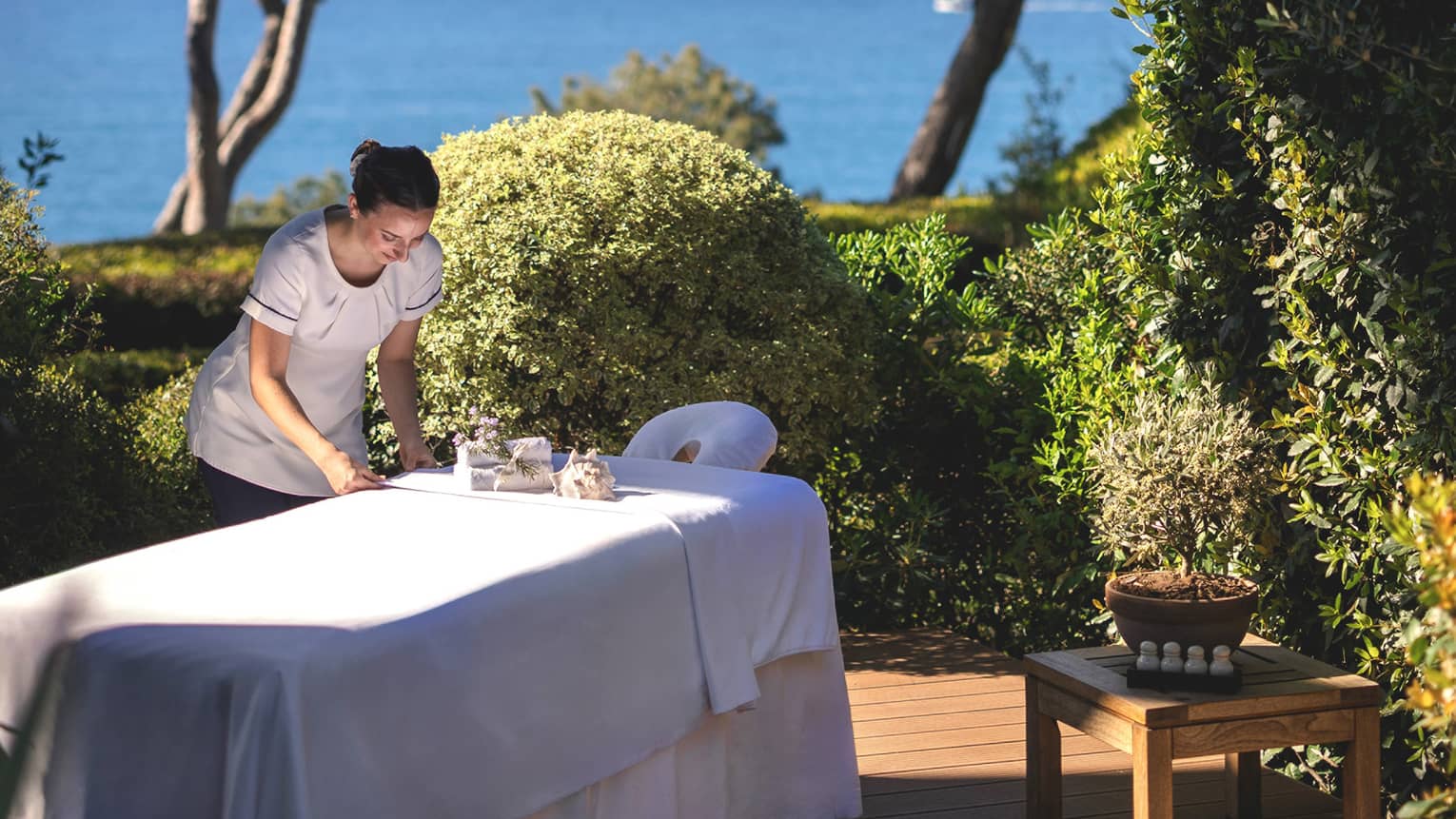 Spa therapist setting up spa table on outdoor platform surrounded by bushes and trees and overlooking the sea
