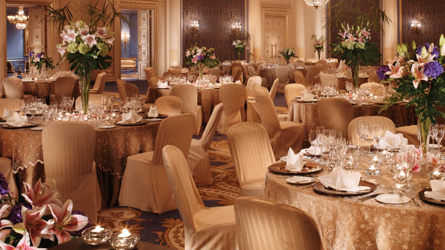 Elegantly set tables with gold tablecloths, flowers in ballroom with textured blue panels chandeliers 