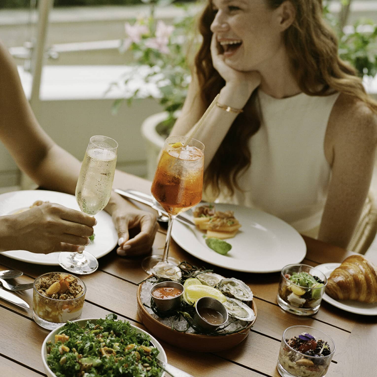 Laughing guests enjoying cold beverages, raw oysters with sauces, smoked salmon, a kale salad, muesli and croissants.